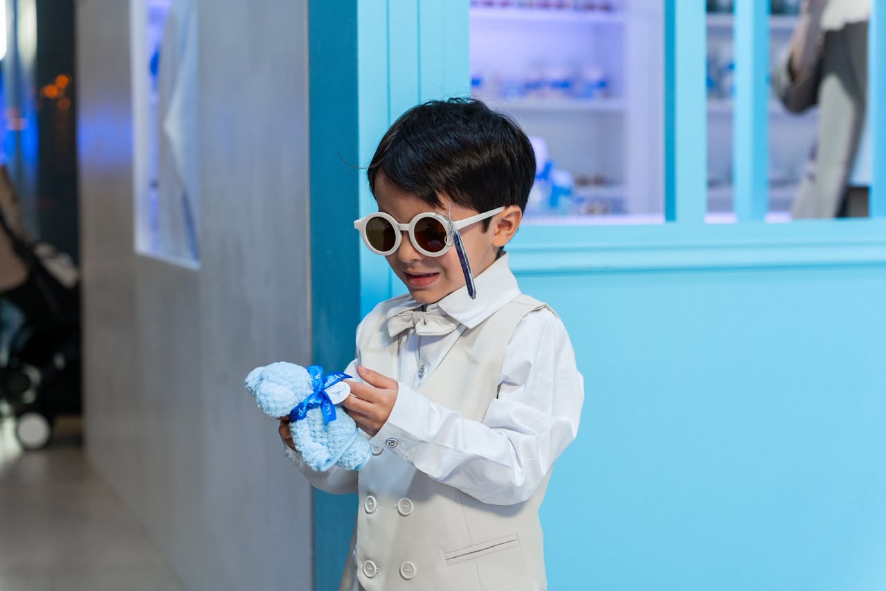 Niño con lentes y traje claro sosteniendo un souvenir de oso azul